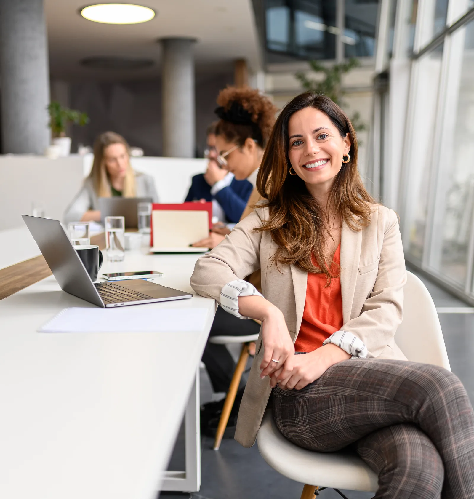 Consultant smiling at her desk while colleagues review documents at a table behind her